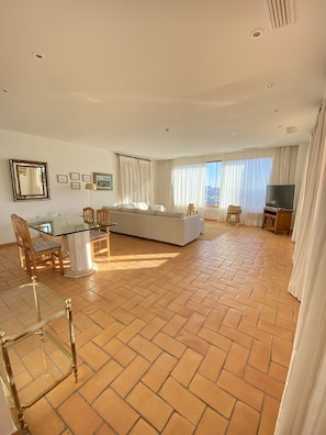 A spacious living area with a sofa, dining table, and chairs. There is a television and a large window with curtains.