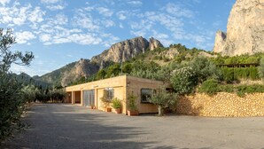 A building with a stone wall and a paved driveway, surrounded by greenery and mountains.