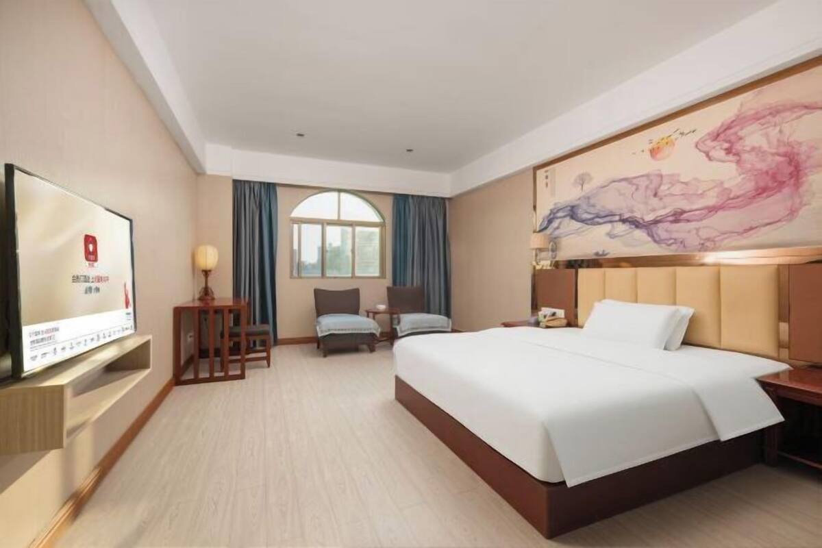 Una habitación de hotel con cama, televisor, escritorio, silla, ventana y un cuadro en la pared.