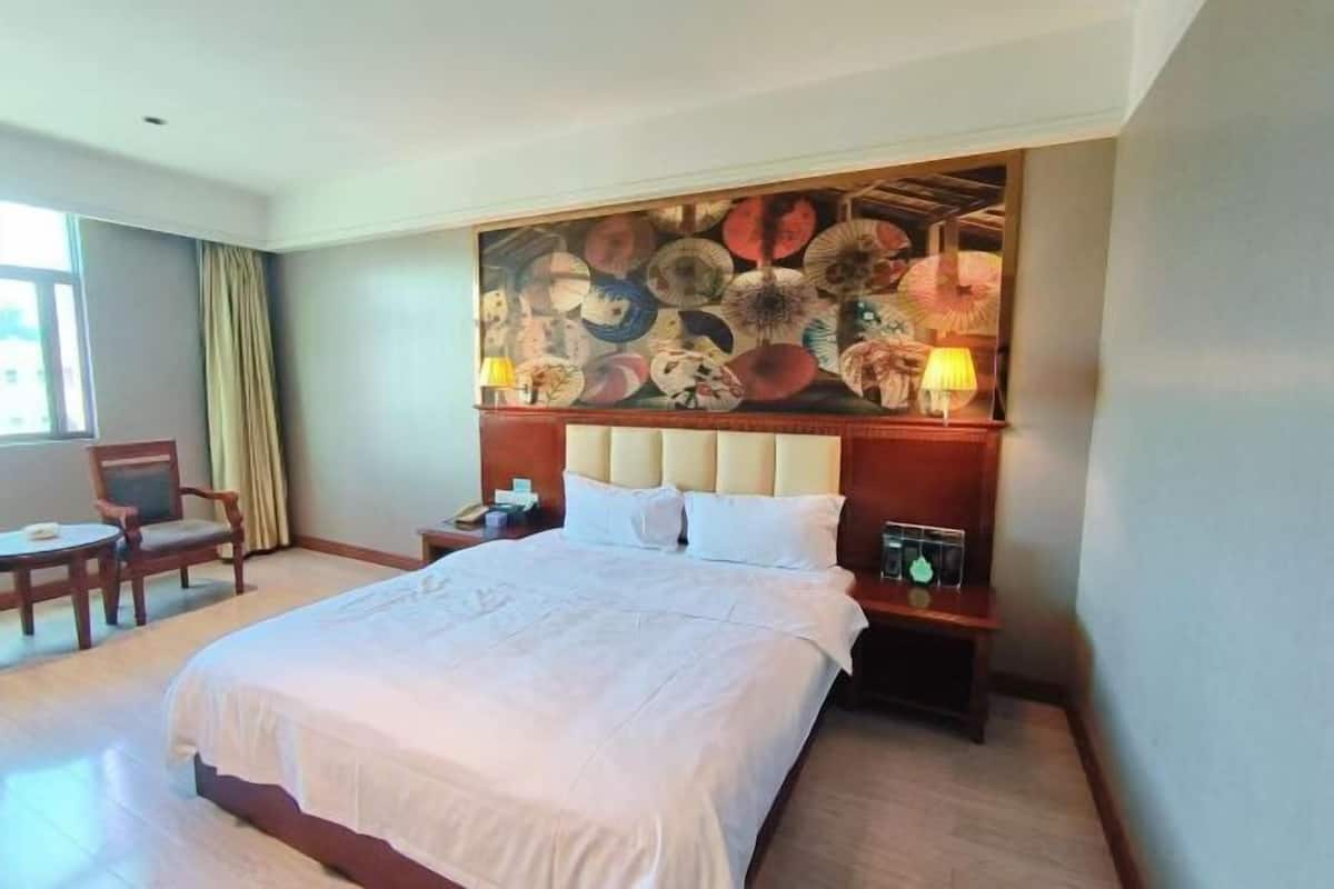 Habitación de hotel con una cama grande, cabecera con un mural floral, mesita de noche con lámpara y una silla.