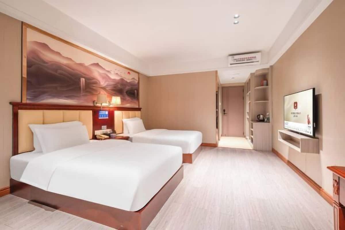 Habitación de hotel con dos camas, un cuadro grande en la pared, un televisor montado y un pequeño escritorio con una lámpara.