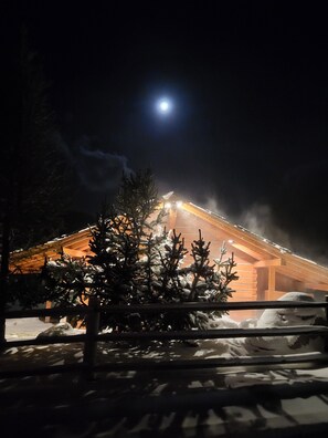A snow-covered landscape with a wooden cabin illuminated by lights, a snow-covered ground, and a tree in the foreground.
