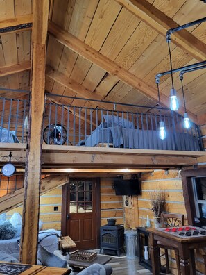 A wooden cabin interior with a loft bedroom, a fireplace, and a dining area.