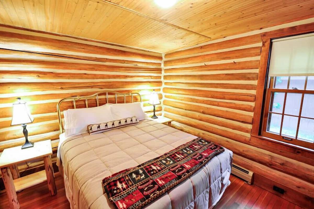 A log cabin room with a bed, a nightstand, a lamp, and a window.