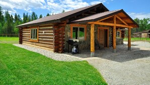 A log cabin with a covered porch, a grill outside, and a gravel driveway. - Glacier Homestead (Coram)
