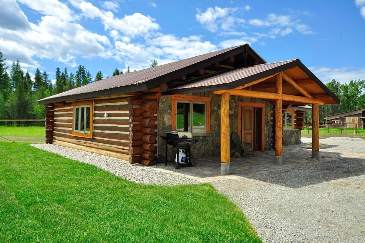 A log cabin with a covered porch, a grill outside, and a gravel driveway.