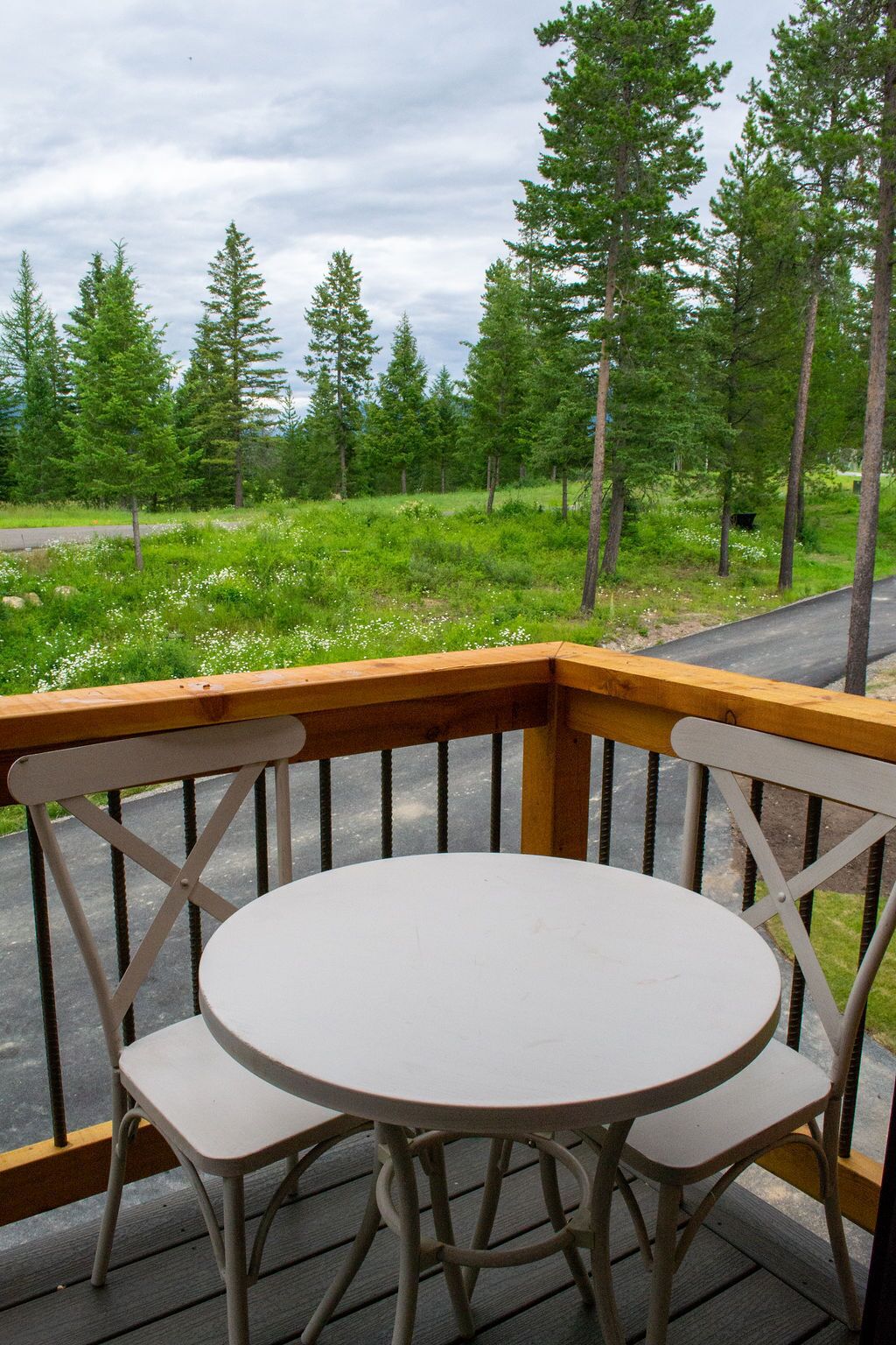 A wooden deck with a round table and two chairs, overlooking a forested area with a road in the background.