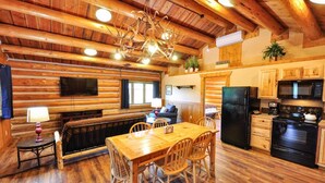 A log cabin interior with a wooden dining table, chairs, a kitchen area with appliances, and a fireplace. - Glacier Homestead (Coram)