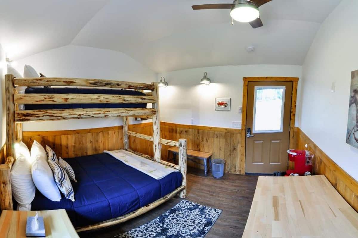 A bunk bed with a wooden frame and blue bedding, a small table with a book, a patterned rug, and a ceiling fan.