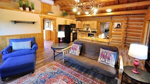A log cabin interior with a living room featuring a blue armchair, a sofa with decorative pillows, a patterned rug, and a chandelier. - Glacier Homestead (Coram)