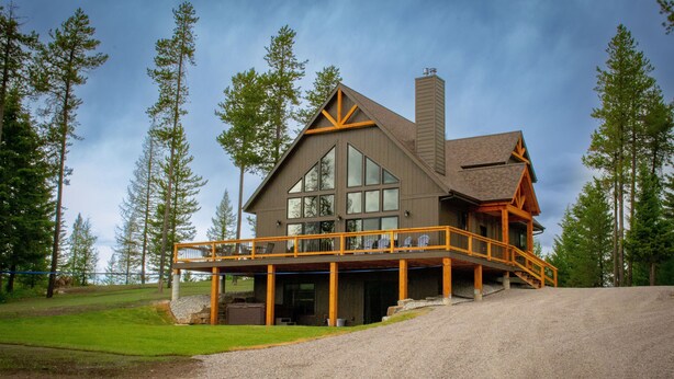 A large, two-story lodge with a prominent balcony and a chimney, surrounded by tall trees. - Glacier Homestead (Coram)