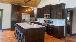 A spacious kitchen with dark wood cabinets, stainless steel appliances, and a large island with bar stools. - Glacier Homestead (Coram)