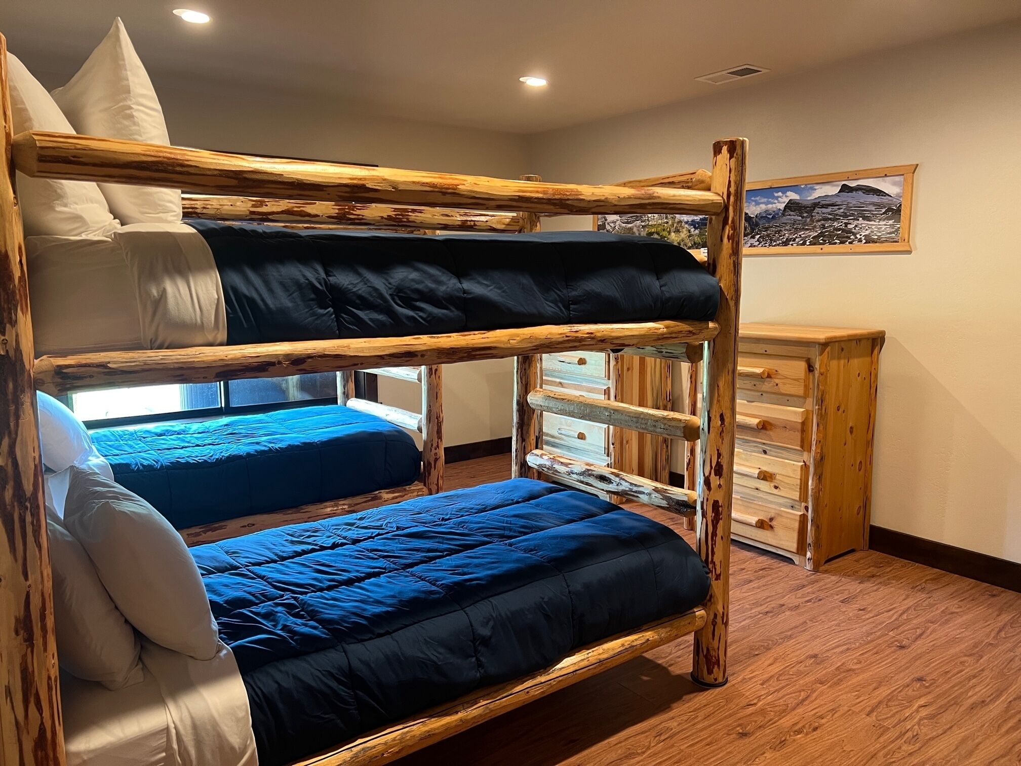 Une chambre avec un lit superposé en bois, deux lits avec une literie bleue, une commode en bois et un tableau encadré au mur.