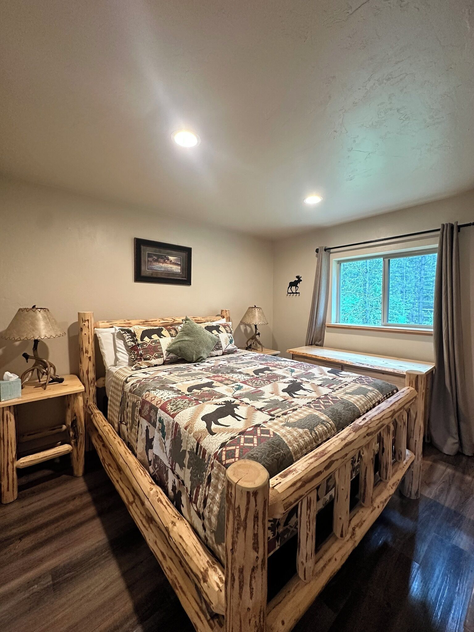 A wooden bed with a patterned quilt, two bedside tables with lamps, a framed picture on the wall, and a window with curtains.