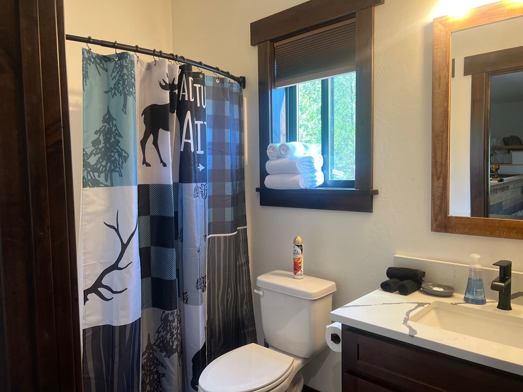 Bagno con tenda della doccia decorata con un motivo di alberi e animali, un water, un lavandino con articoli da toilette e una finestra con asciugamani.