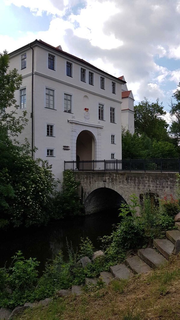En vit byggnad med en central valvöppning, en stenbro och frodig grönska.