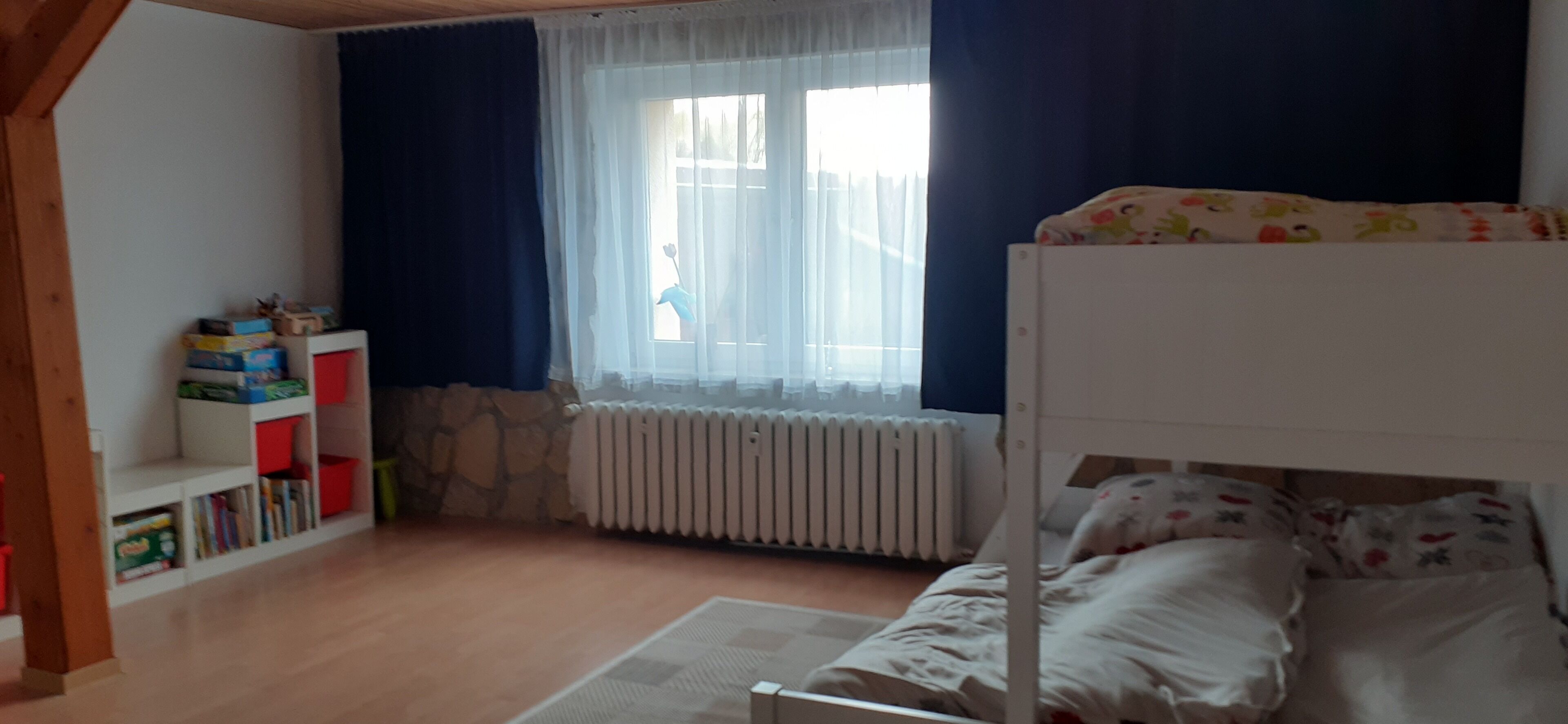Ein Kinderzimmer mit Etagenbett, Heizkörper, einem Fenster mit Vorhängen und einem Bücherregal mit Spielzeug.