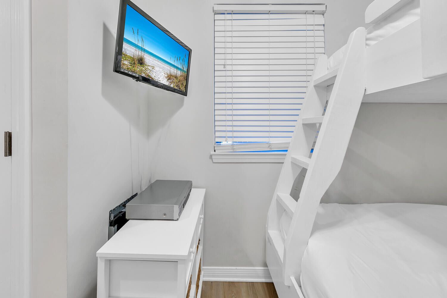 A bedroom with a bunk bed, a flat-screen TV mounted on the wall, a small white table, and a window with blinds.