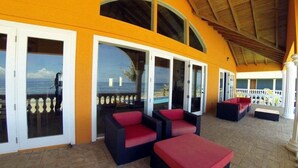 An outdoor lounge area with wicker furniture and red cushions, large glass doors leading to a balcony with a view of the sea. - Casa Naranja - Upscale Living with a Private Pool (Utila)