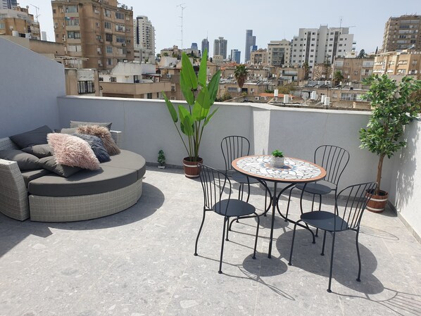 A rooftop terrace with a round table and chairs, a sofa with cushions, and potted plants.