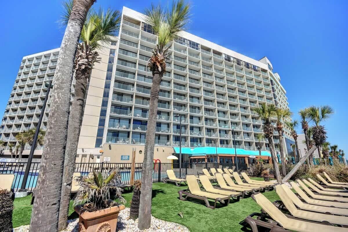 A large hotel with multiple floors, a pool area with lounge chairs, and palm trees.