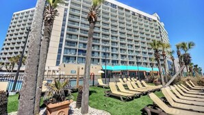 A large hotel with multiple floors, a pool area with lounge chairs, and palm trees.
