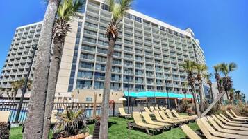 A large hotel with multiple floors, a pool area with lounge chairs, and palm trees.