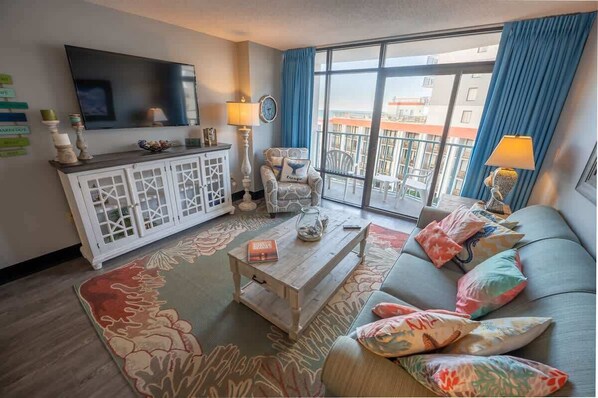 A modern living room with a flat-screen TV, a white cabinet with glass doors, a patterned rug, a sofa with colorful cushions, and a balcony with blue curtains.