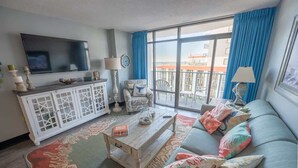 Ein Wohnzimmer mit einer Couch, einem Couchtisch, einem Fernseher und einem Balkon mit blauen Vorhängen.