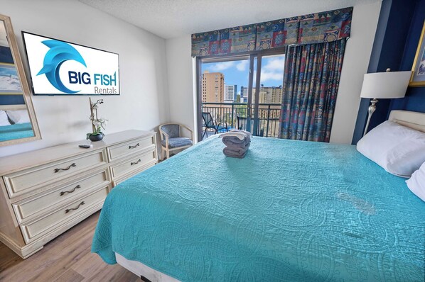 A hotel room with a large bed, a dresser, a TV displaying 'BIG FISH Rentals', a balcony with a view of buildings, and a patterned curtain.