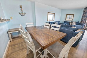 A dining area with a wooden table and white chairs, a blue sofa with patterned cushions, and a wall-mounted anchor decoration.