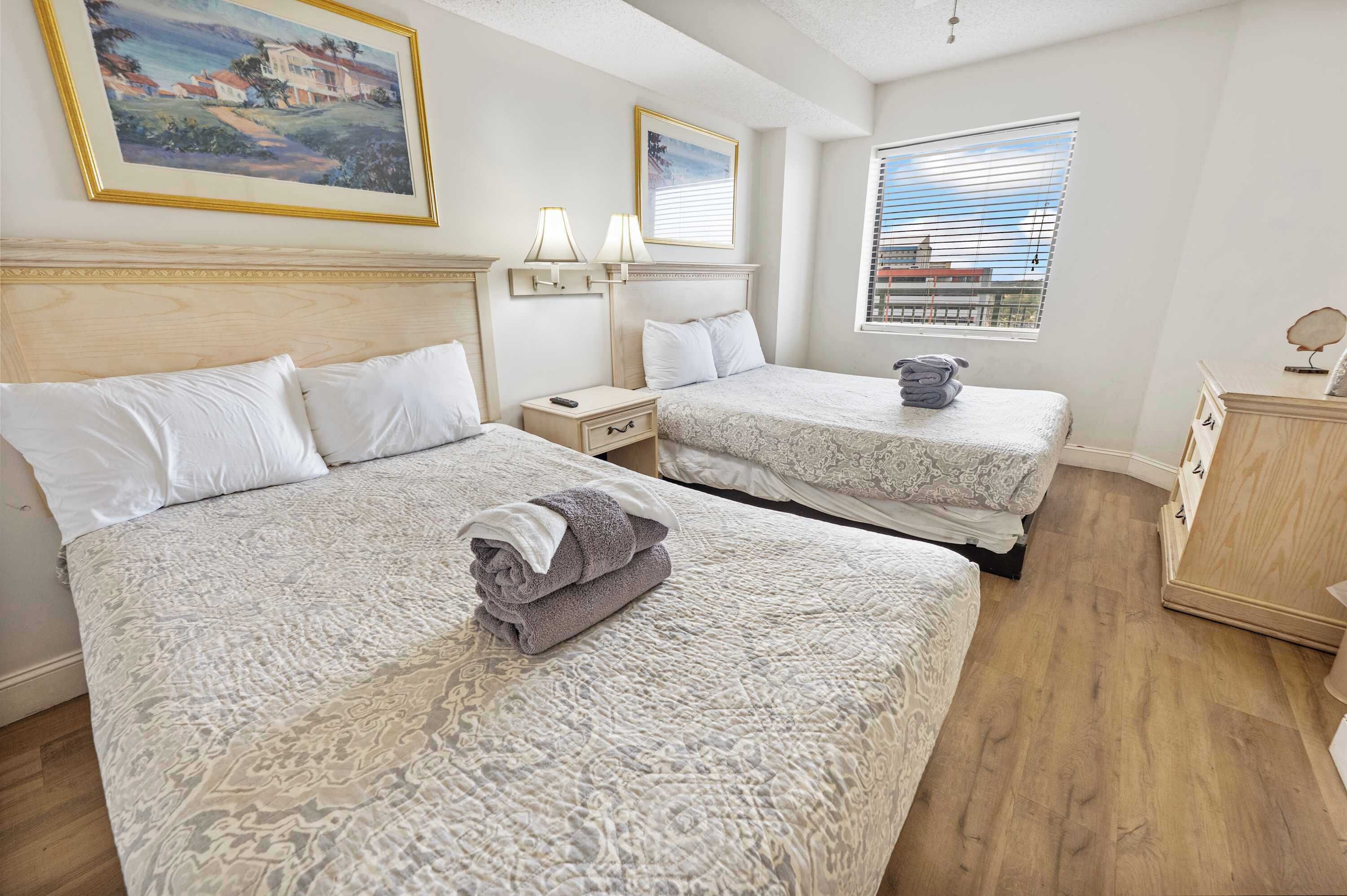 Une chambre d’hôtel avec deux lits, une table de chevet, une lampe, une fenêtre avec des stores et un tableau au mur.