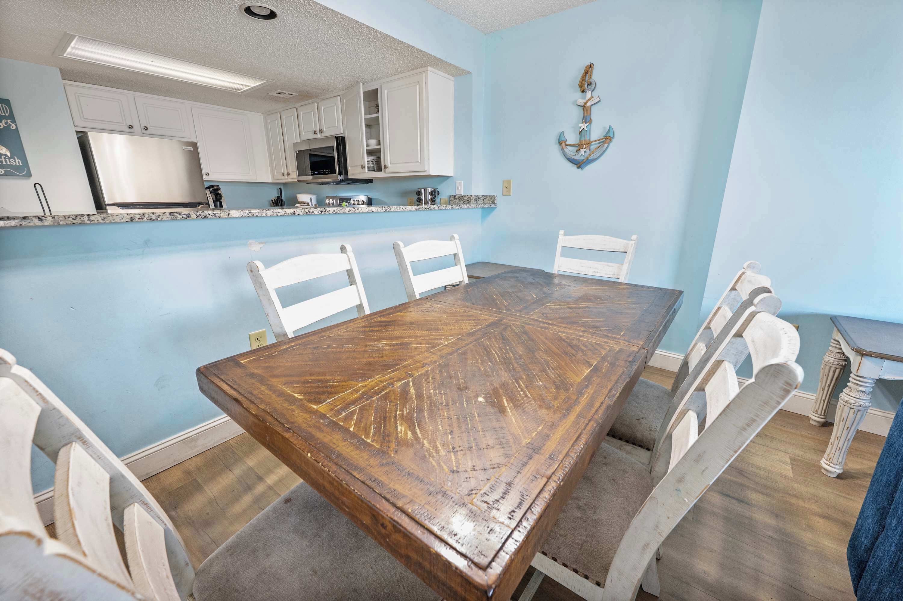 A dining area with a wooden table and white chairs, a kitchen with white cabinets, and a blue accent wall with an anchor decoration.