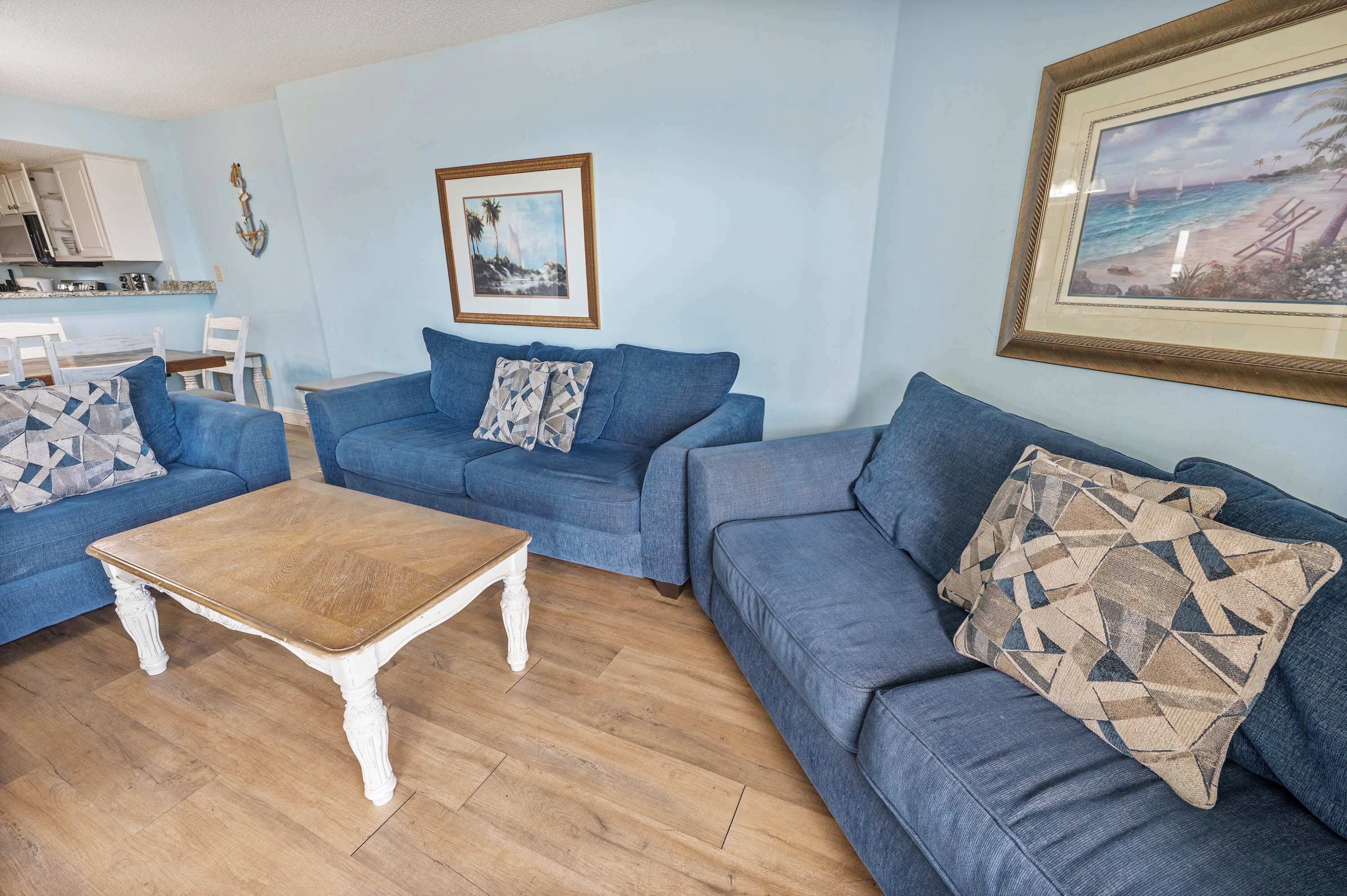 A living room with a blue sofa set, a wooden coffee table, and a framed beach painting.