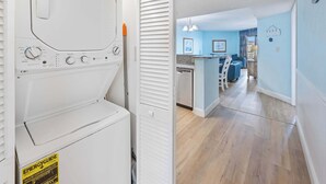A laundry area with a front-loading washer and dryer, a white door, and a hallway leading to a kitchen area with blue walls and wooden flooring.