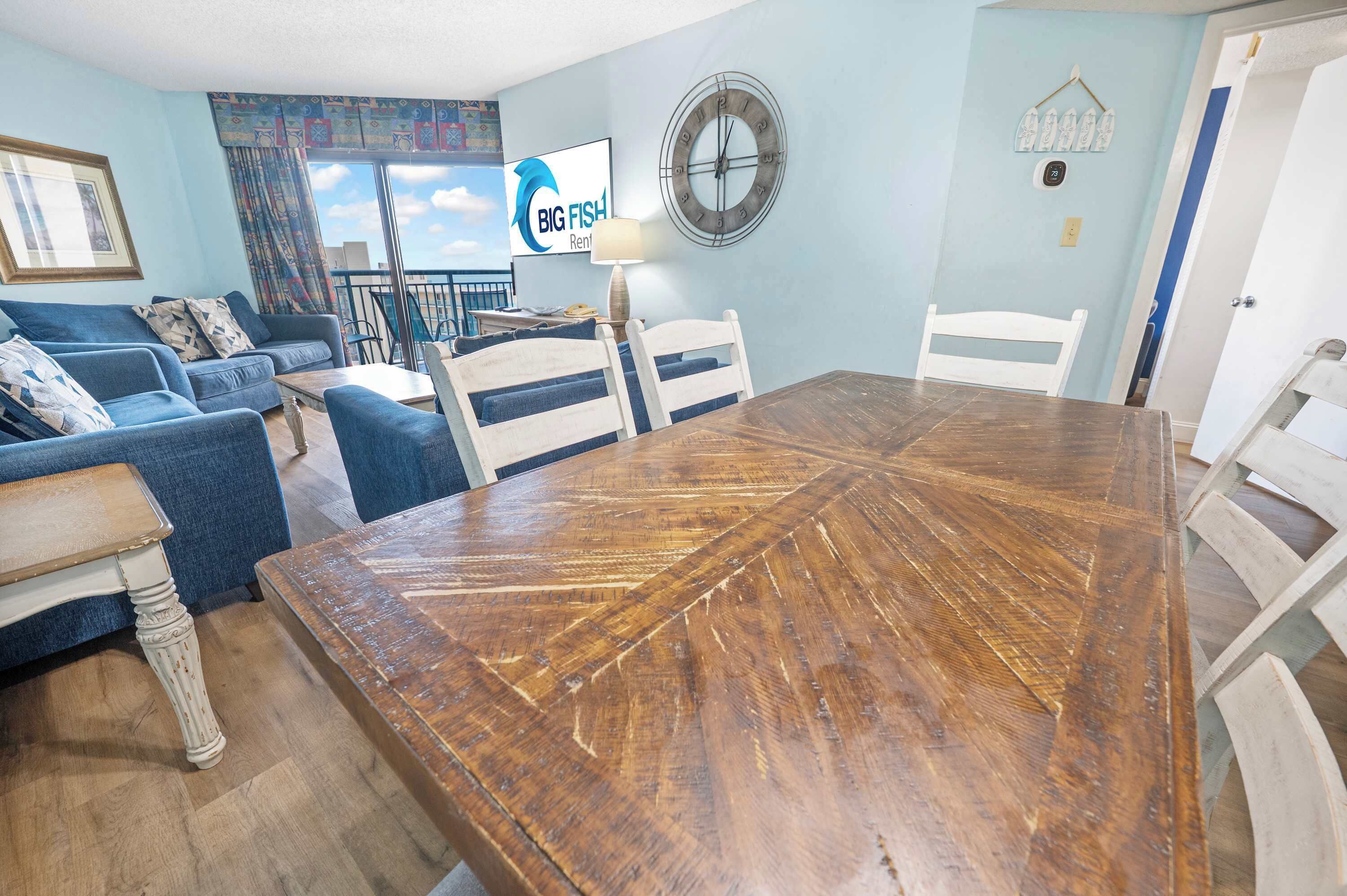 A living room with a wooden dining table, blue sofas, and a large window with a view.