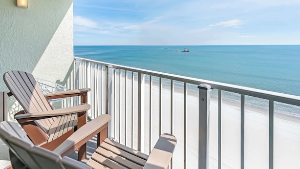 A balcony with two Adirondack chairs and a small table, overlooking a beach with clear blue water and a distant boat. - New Direct Ocean View, Exclusive Sand Beach Club! (Myrtle Beach)