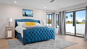 Un dormitorio con una cama tapizada en azul, vistas al exterior y un cuadro en la pared.