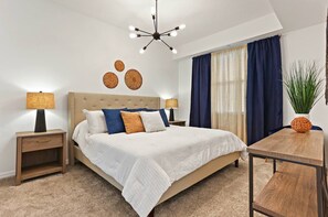 A bedroom with a large bed, two bedside tables, a wooden nightstand, a ceiling light fixture, a window with curtains, and a small table with a plant.