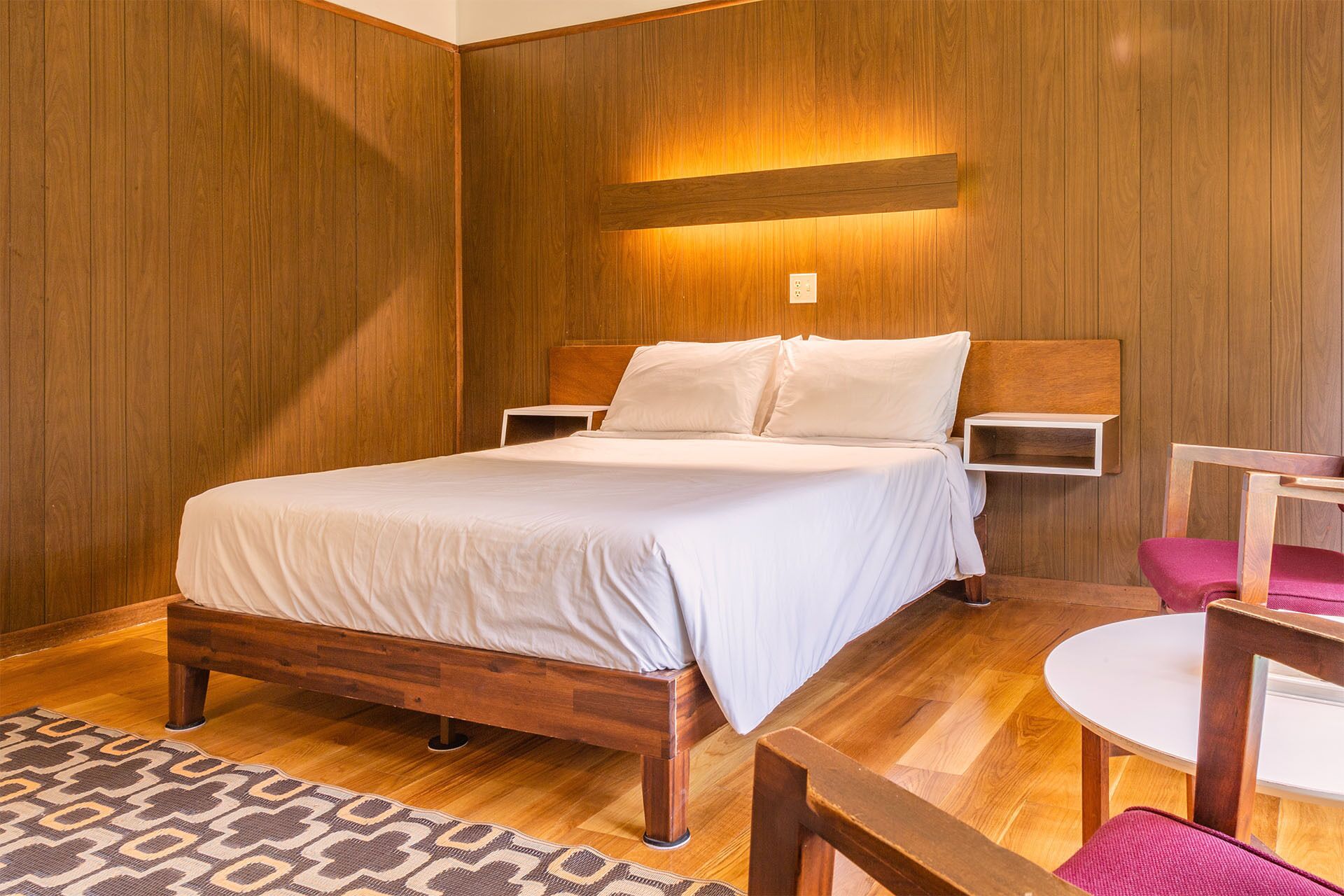 Una habitación con paneles de madera, una cama, mesitas de noche, una silla y una pequeña mesa.