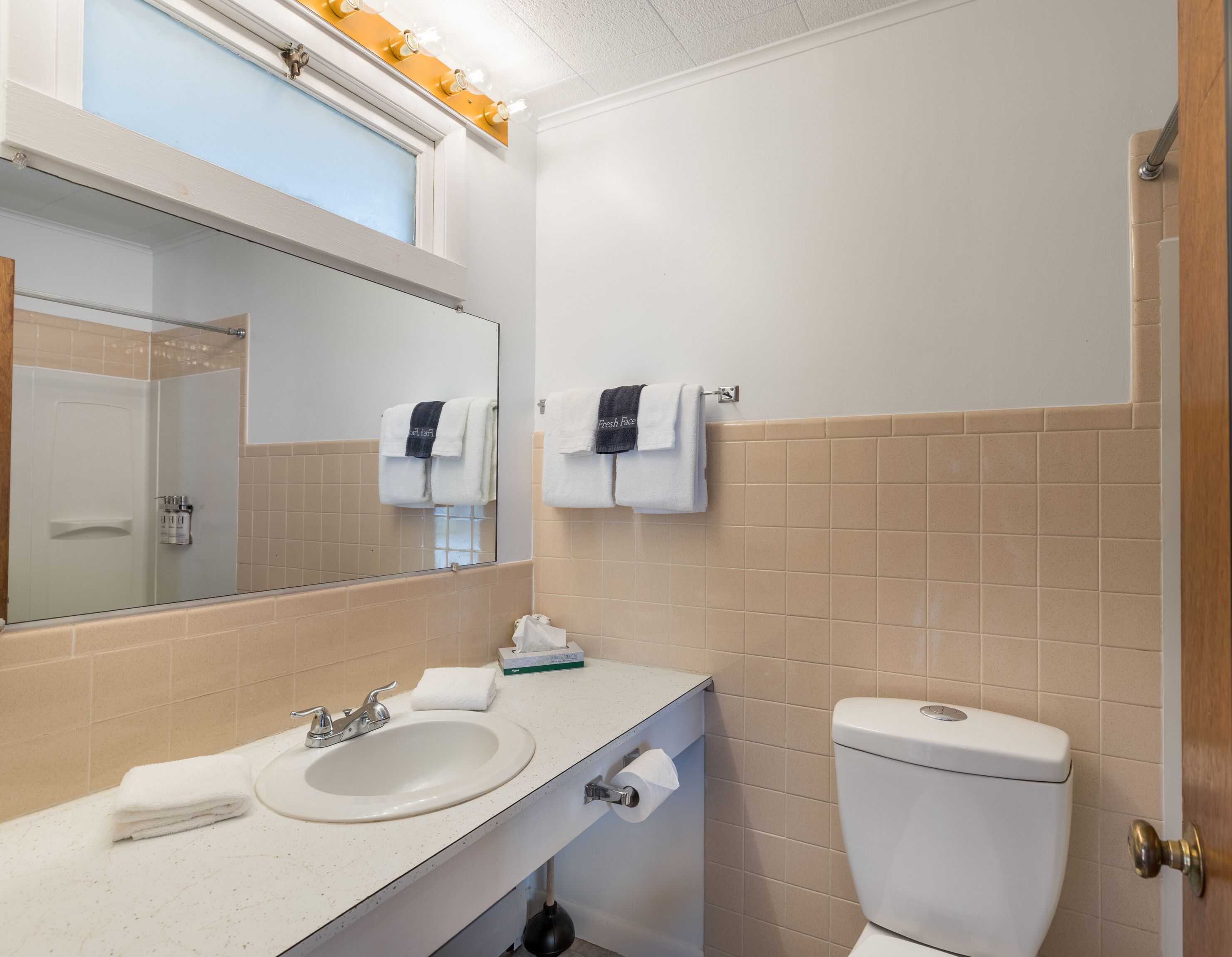 A bathroom with a sink, mirror, towel rack, toilet, and a window with light fixtures.