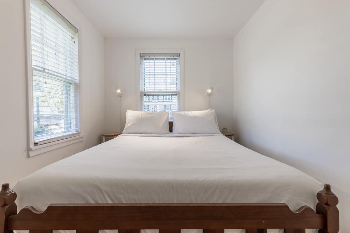 Une chambre à coucher avec un lit en bois, une literie blanche, deux tables de chevet avec des lampes et une fenêtre avec des stores.