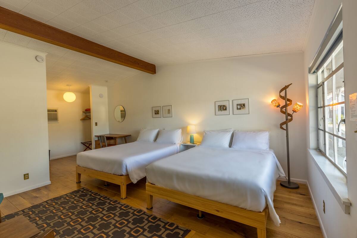 Une chambre avec deux lits, un plancher en bois, un tapis à motifs, une lampe sur pied et une fenêtre donnant sur un paysage agréable.