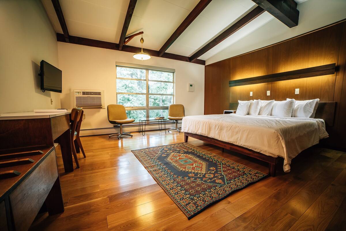 Une chambre avec un grand lit, un plancher en bois, un bureau avec une chaise, un téléviseur fixé au mur et une fenêtre donnant sur des arbres.