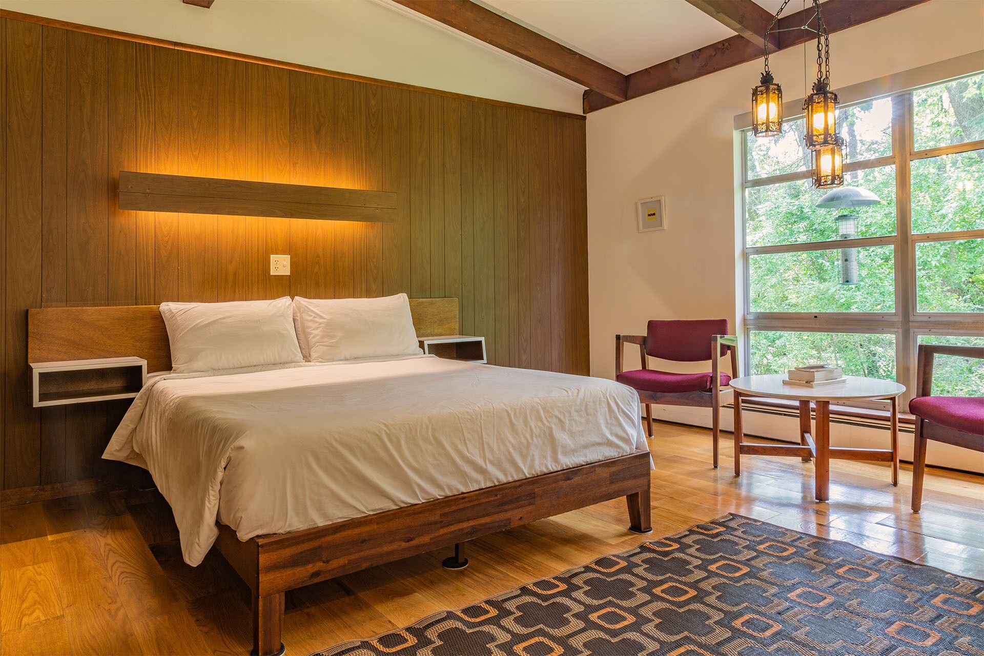 A bedroom with a wooden bed, a nightstand, a chair, a small table, and a window with a view of greenery.