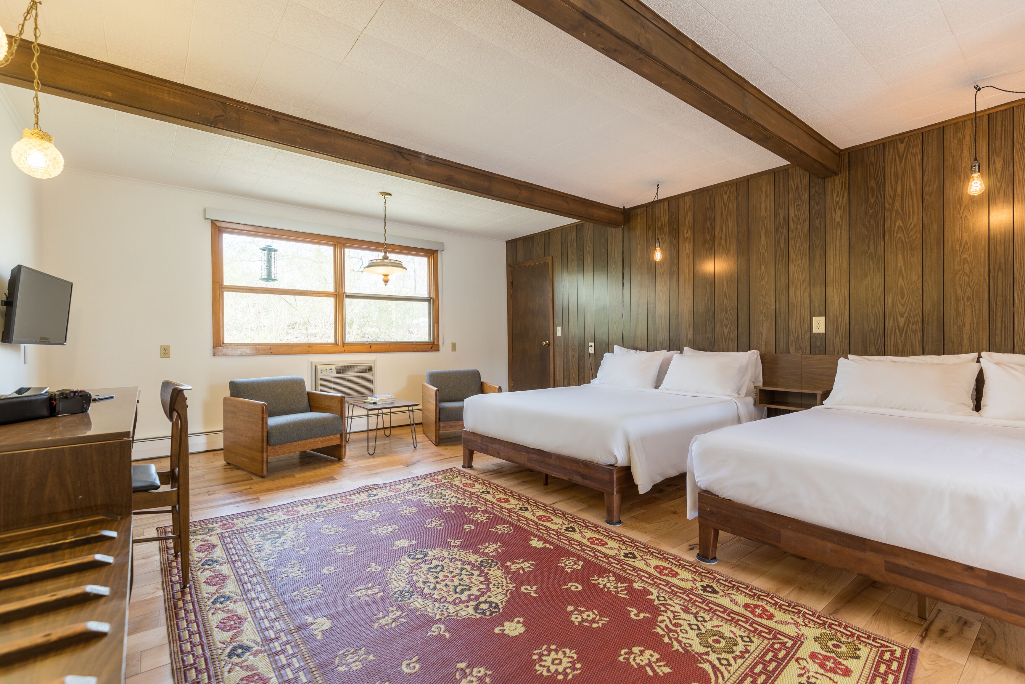 A hotel room with two beds, a wooden floor, a red patterned rug, a desk with a chair, a television, and a window with a view of trees.