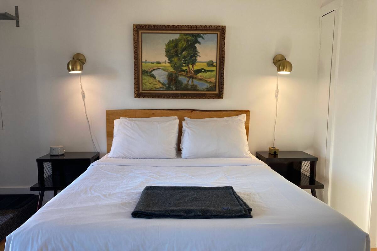 Un lit bien fait, avec du linge de lit blanc, deux tables de chevet avec des lampes et un tableau représentant un paysage, encadré, accroché au mur.
