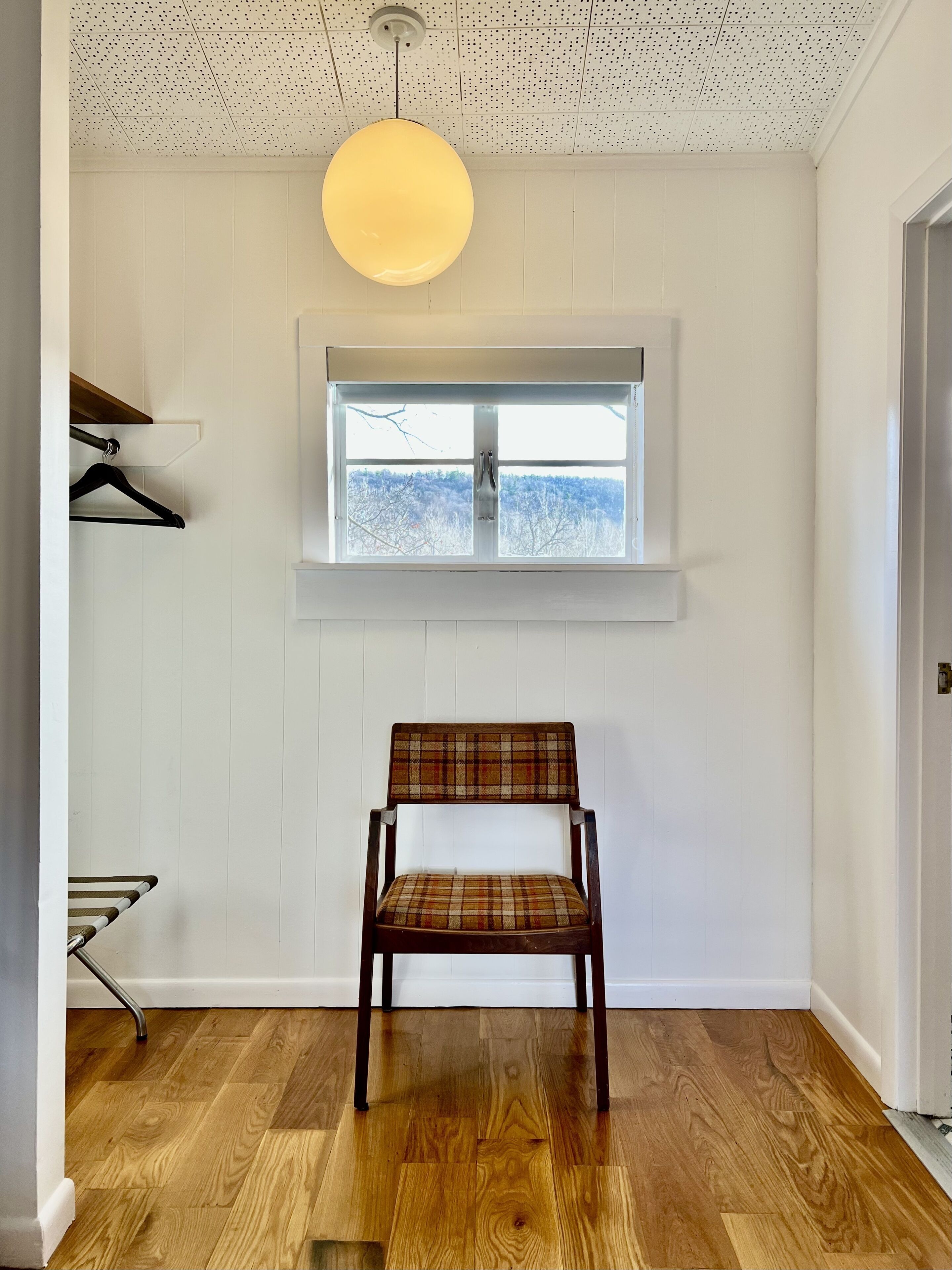 Una habitación con suelo de madera, una silla estampada de tartán, una lámpara colgante, una ventana con vistas a árboles y un perchero.