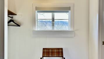 Una habitación con suelo de madera, una silla estampada de tartán, una lámpara colgante, una ventana con vistas a árboles y un perchero.