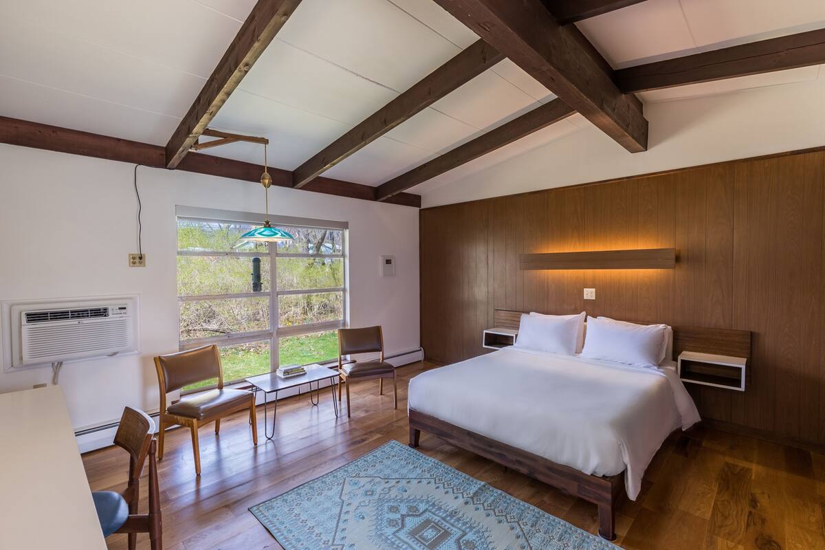 Une chambre avec un grand lit, des poutres apparentes en bois au plafond, une fenêtre avec vue, un bureau avec des chaises et une table de chevet.
